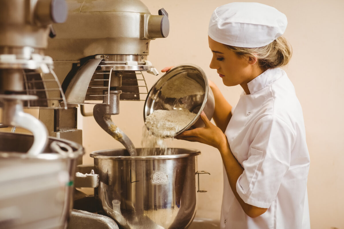 A baker adds flour to a large mixing bowl, creating goods in bulk.