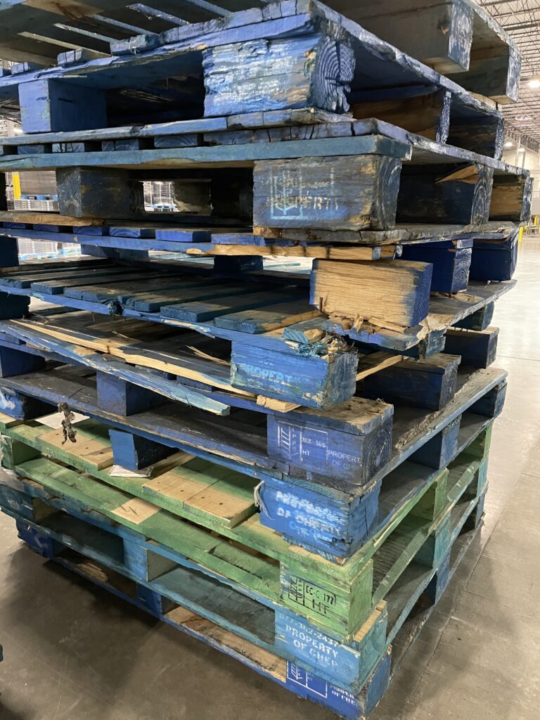 A close-up view of disproportionately sized wooden pallets stacked in a food manufacturing environment reveals extreme signs of wear, including chipping and breakage.