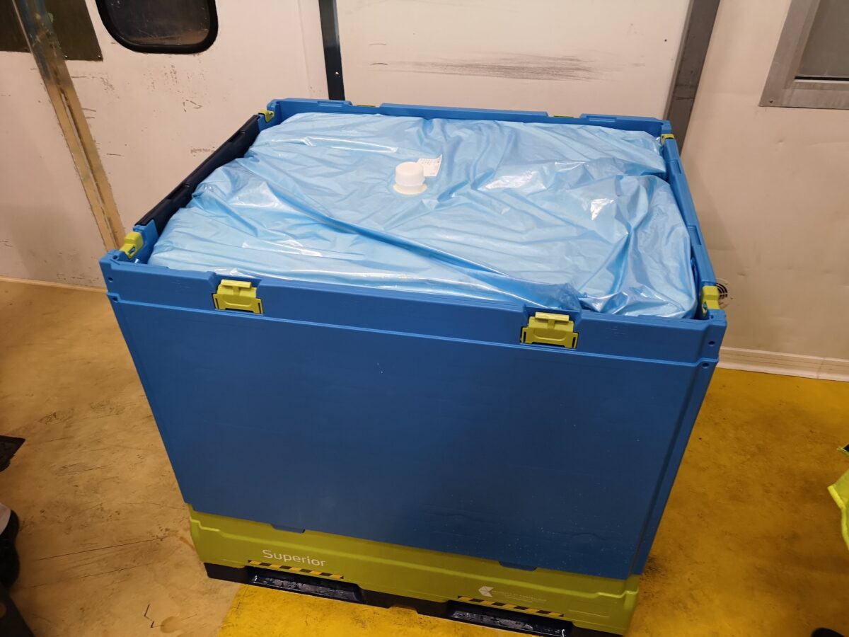 A Tosca Superior Hybrid bin with a liner, filled and sealed, sits in a manufacturing facility. This food-grade IBC tote is designed to provide better product protection for the food packaging industry.