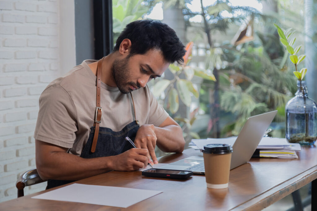 Indian man sits to calculate income and expenses accounts. Man employee wearing apron sitting on table and clear account before closing shop, personal business concept.